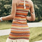 Colorful Aztec print halter neck mini dress with fringe hem detail on a woman standing near a white fence in a green field.