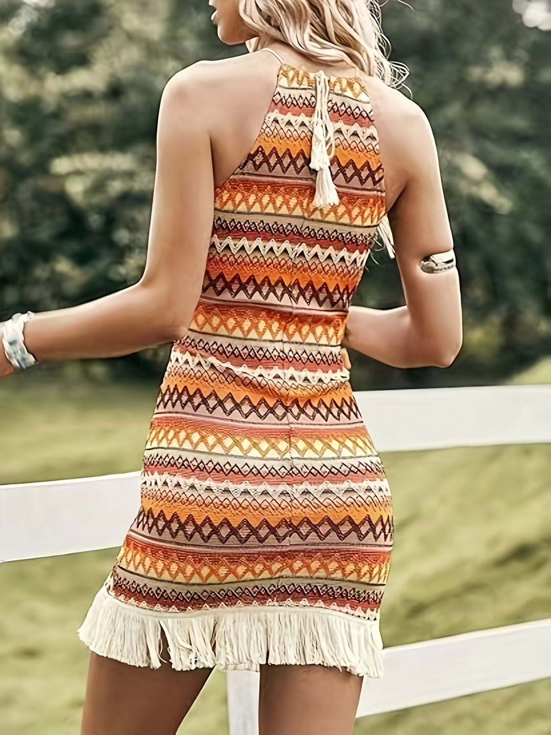 Colorful Aztec print halter neck mini dress with fringe hem detail on a woman standing near a white fence in a green field.