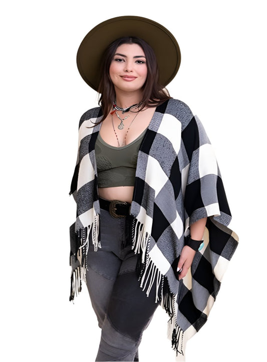 Black and white plaid poncho with fringe, worn over a green top and grey jeans, accessorized with a hat.