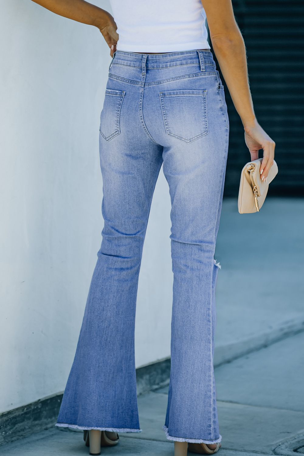 Back view of button-fly distressed raw hem flare jeans in light blue denim with a high waist and frayed hem.