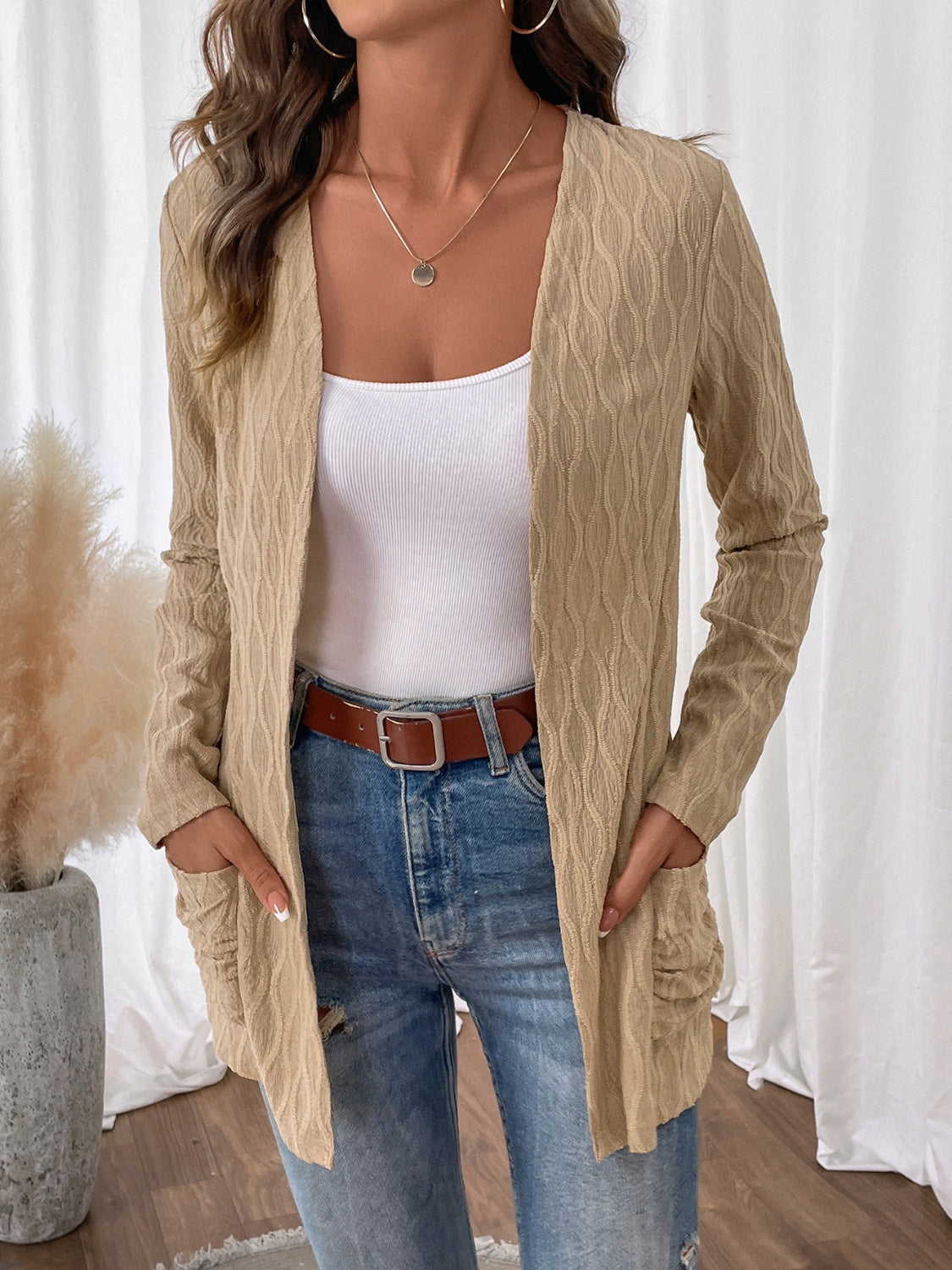 Woman wearing a beige cable knit cardigan with a textured design, styled with a brown belt and denim jeans.