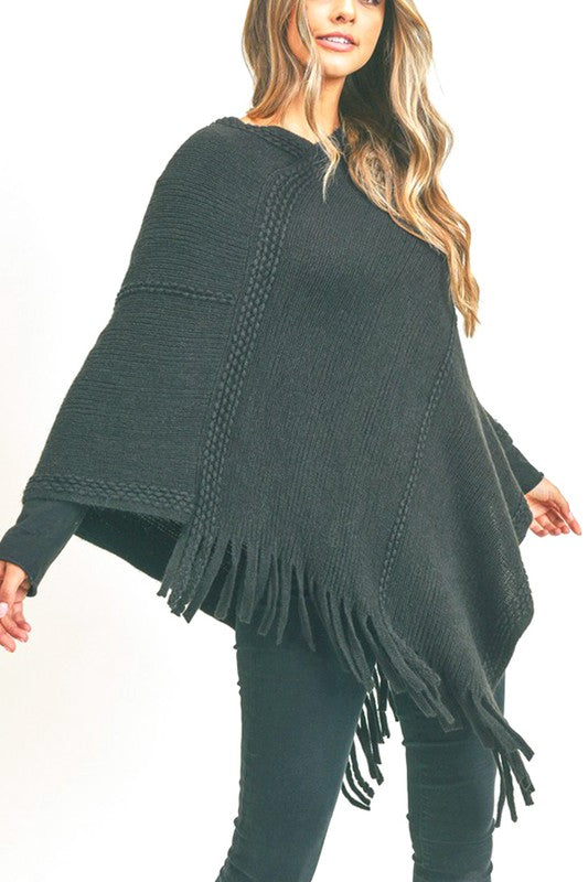 Black knit poncho with fringe detailing, shown layered over a casual outfit, offering a stylish and warm look.