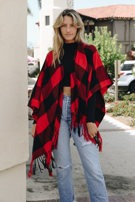 A cozy red and black plaid fringe poncho, draped casually over a black top and jeans.