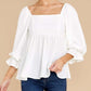 White ruffled babydoll blouse with square neckline, styled with jeans.
